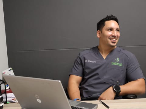 Dr BD Haupt in HealthPLUS scrubs at a clinic desk with laptop