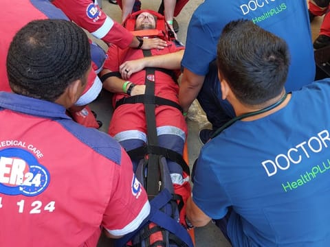 HealthPLUS doctors in blue scrubs and ER24 paramedics in red uniforms stabilising a patient on a spinal board at a sporting event